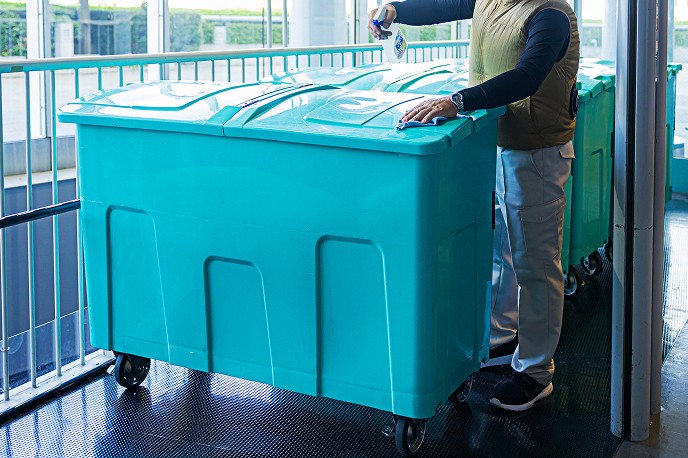 スタッフがスプレーとウエスを使用し、大型の廃棄物回収用コンテナ（ゴミ箱）の蓋を丁寧に拭き上げている様子