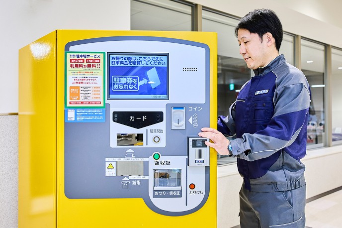黄色い駐車場の料金精算機の前に立ち、パネルを操作して点検を行う作業着姿の男性スタッフ