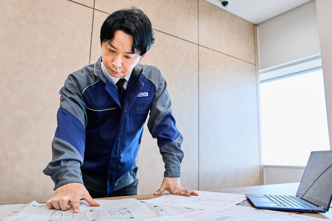作業服を着た男性が、机の上に広げた建築図面を指差して内容を確認している様子