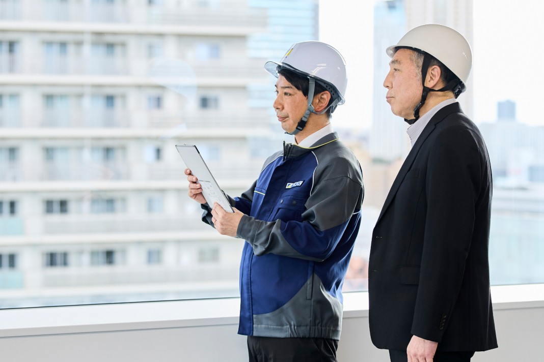 ヘルメットを着用した作業服の担当者とスーツ姿の男性が、窓際で並んで立ち、タブレットを手に外の様子を視察している様子