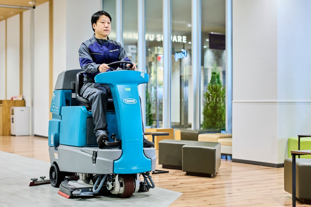 作業服姿のスタッフが、搭乗式の大型床洗浄機を運転し、商業施設のフロアを清掃している様子