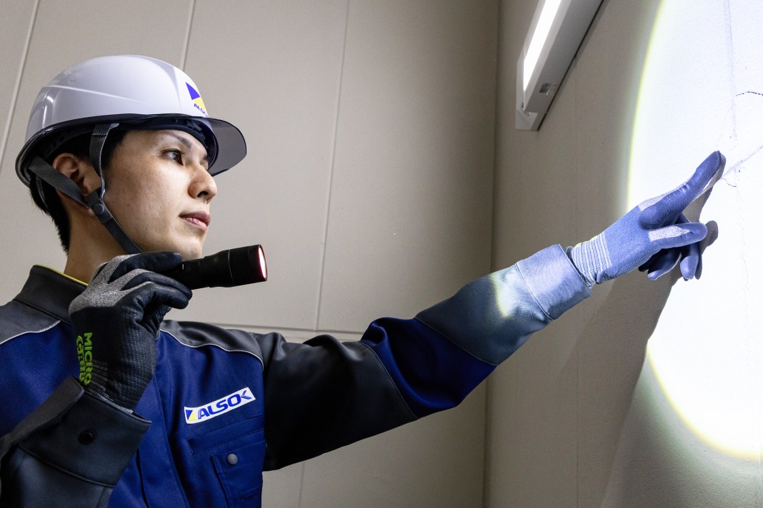定期点検のため、ALSOKの制服とヘルメットを着用した巡回技術者が、ライトで壁を照らし、指差して確認している様子